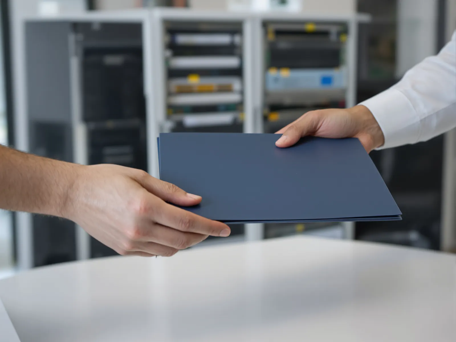 Two pairs of hands passing a closed blue project folder across a desk, with a finished equipment rack out of focus behind