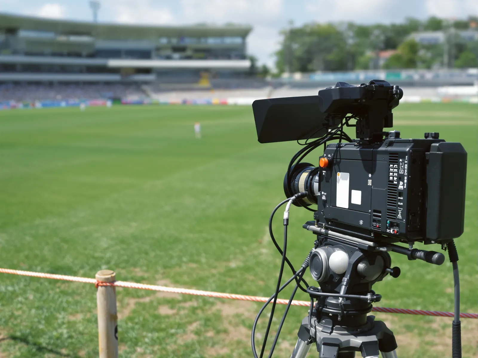 A broadcast television camera on a tripod at the boundary rope of a cricket ground, the stands out of focus behind it